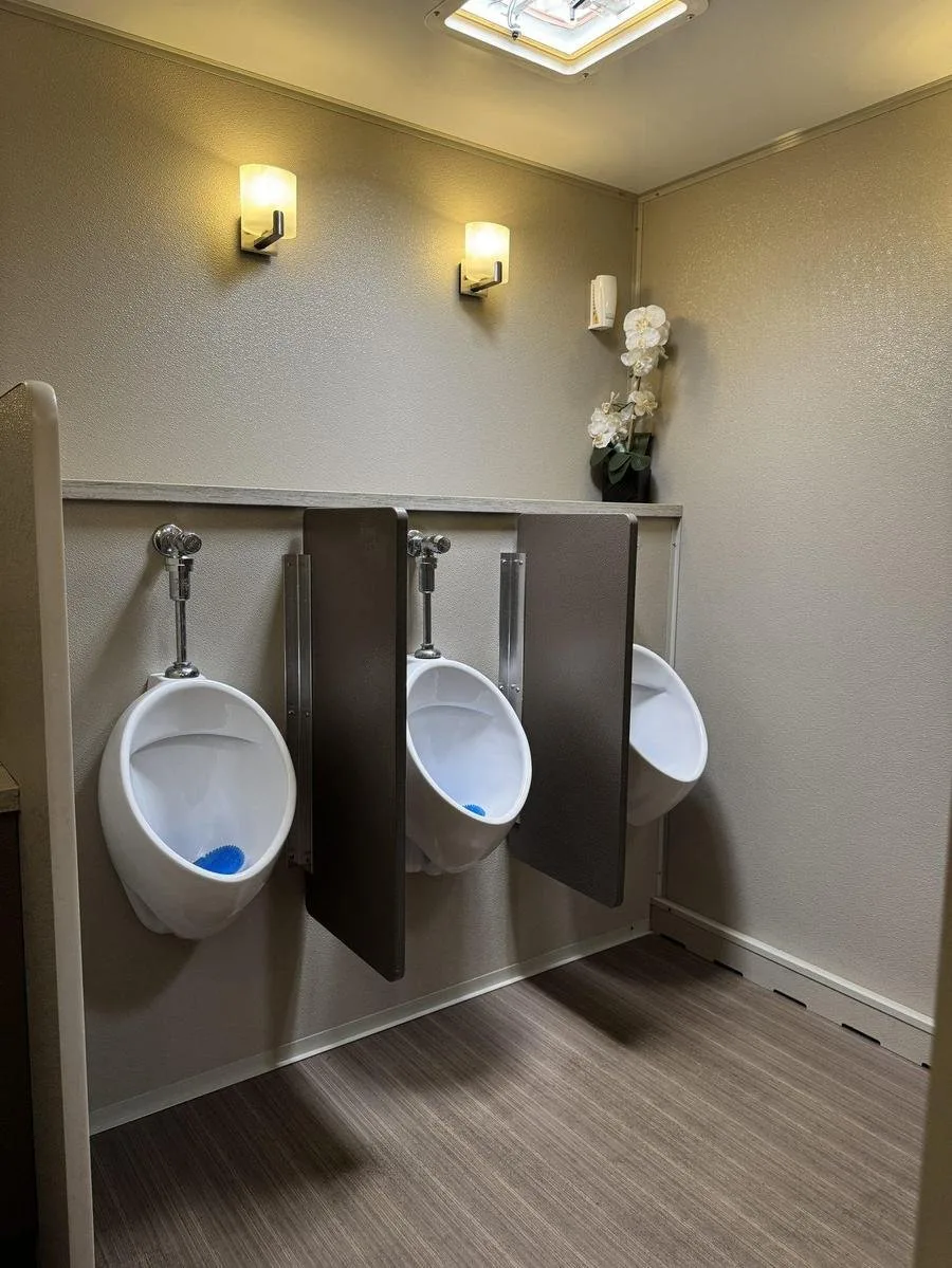 Men's area inside a restroom trailer with urinals, privacy dividers, sconce lighting, and wood-look flooring